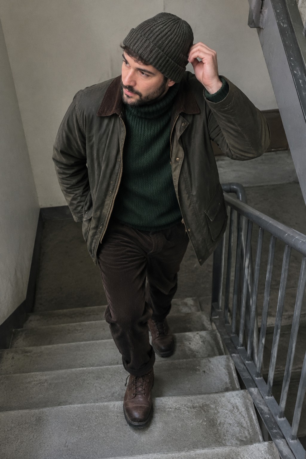 A man wearing a dark green ribbed turtleneck, olive-brown jacket, dark corduroy trousers, brown lace-up boots, and a charcoal beanie as he walks up concrete stairs.