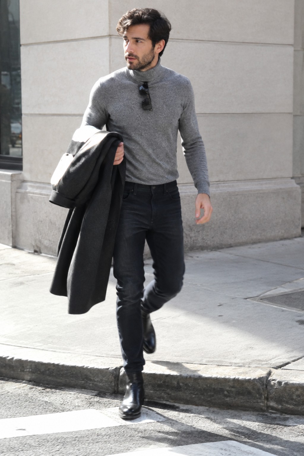 A man in a gray turtleneck, dark jeans, and black leather boots carries a charcoal overcoat and wears sunglasses at his collar.