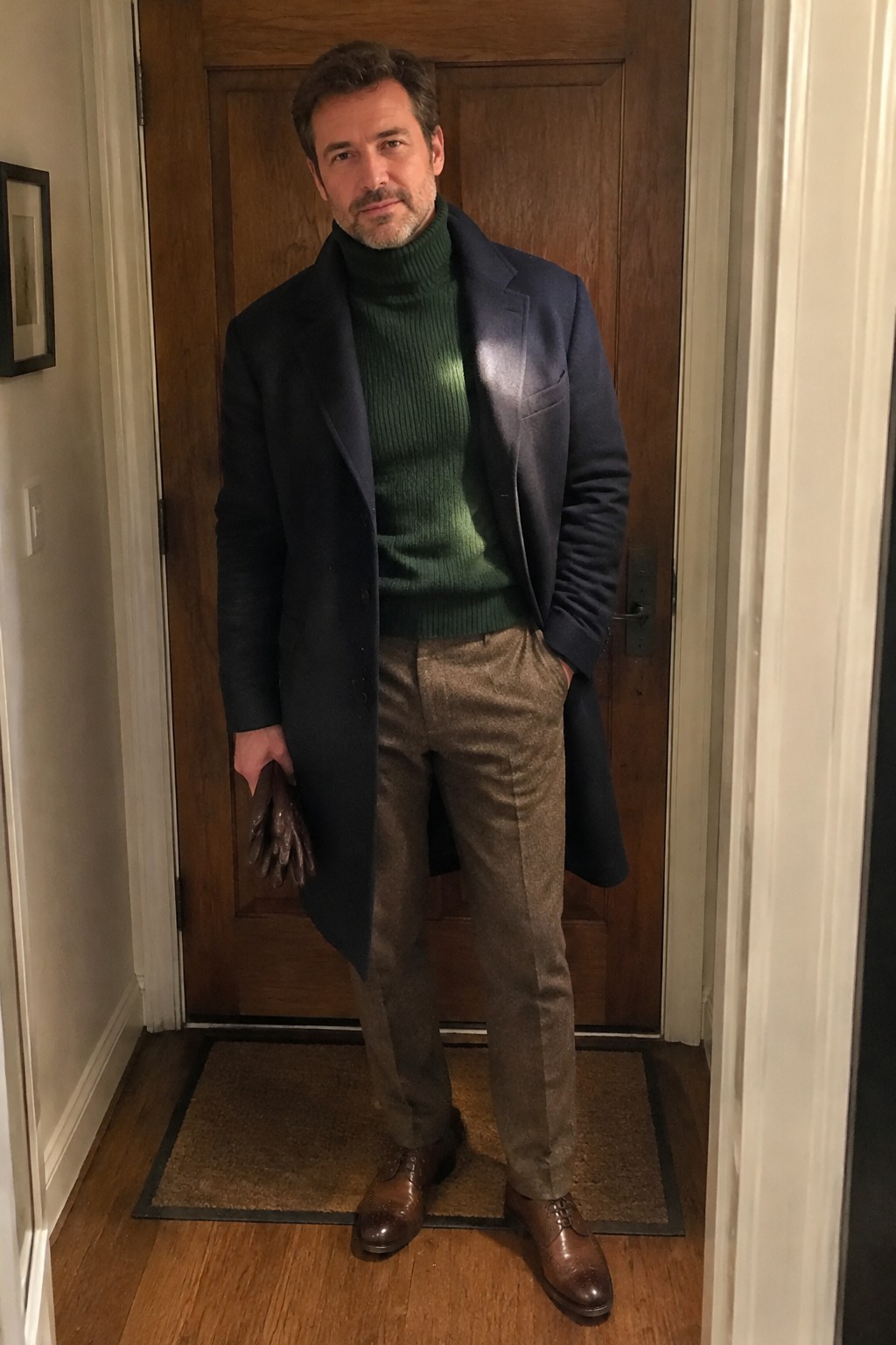 A man wearing a green ribbed turtleneck, long navy overcoat, taupe-brown trousers, brown leather shoes, and gloves.
