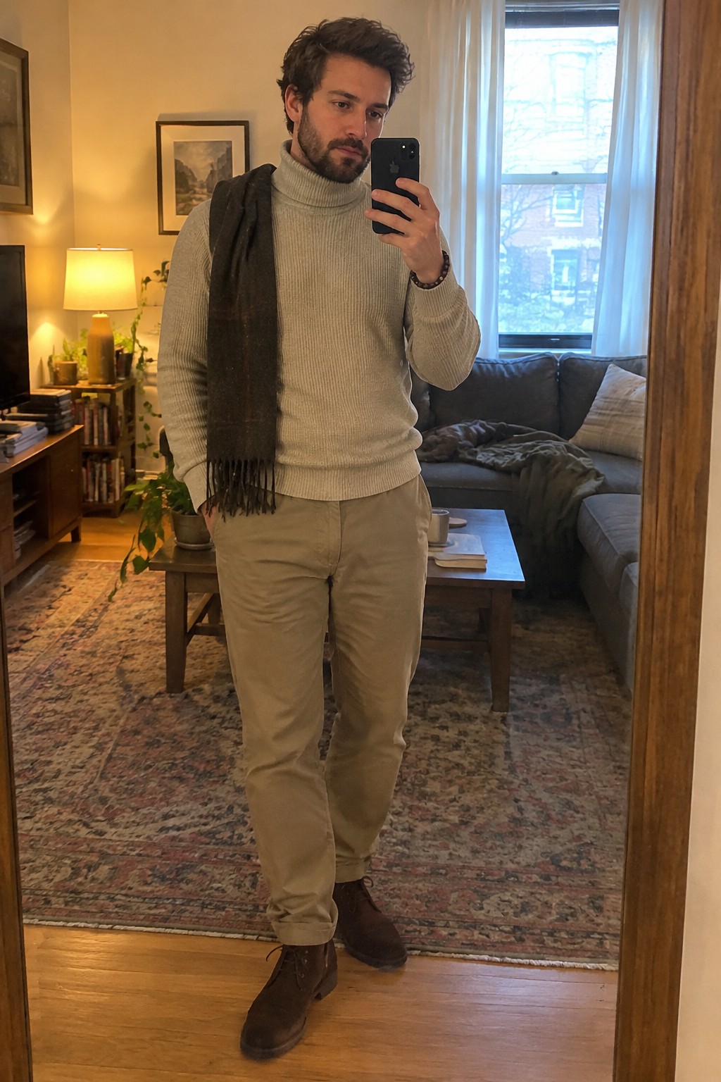 A man wears a light gray ribbed turtleneck, dark brown scarf, tan chinos, brown suede lace-up boots, and a wristwatch.