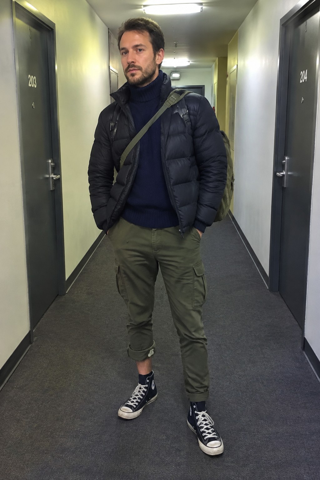 A man wears a navy turtleneck and quilted puffer jacket with olive cargo trousers, black high-top sneakers, and an olive backpack.