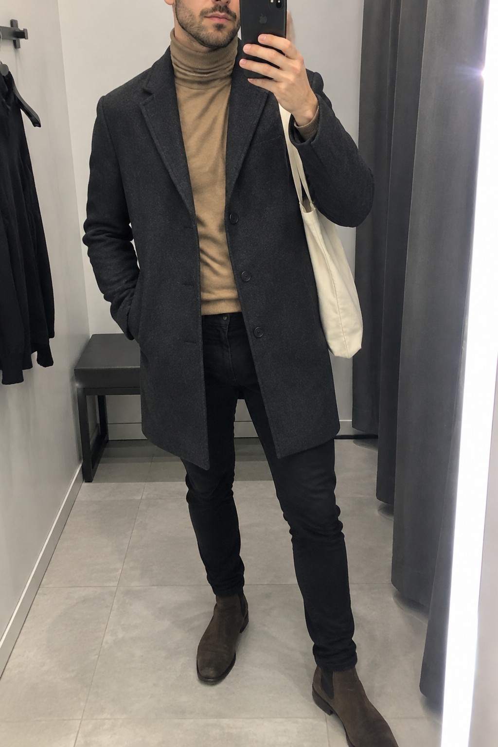 Man wearing a charcoal overcoat over a camel turtleneck with black slim jeans, brown suede Chelsea boots, and a cream tote bag.