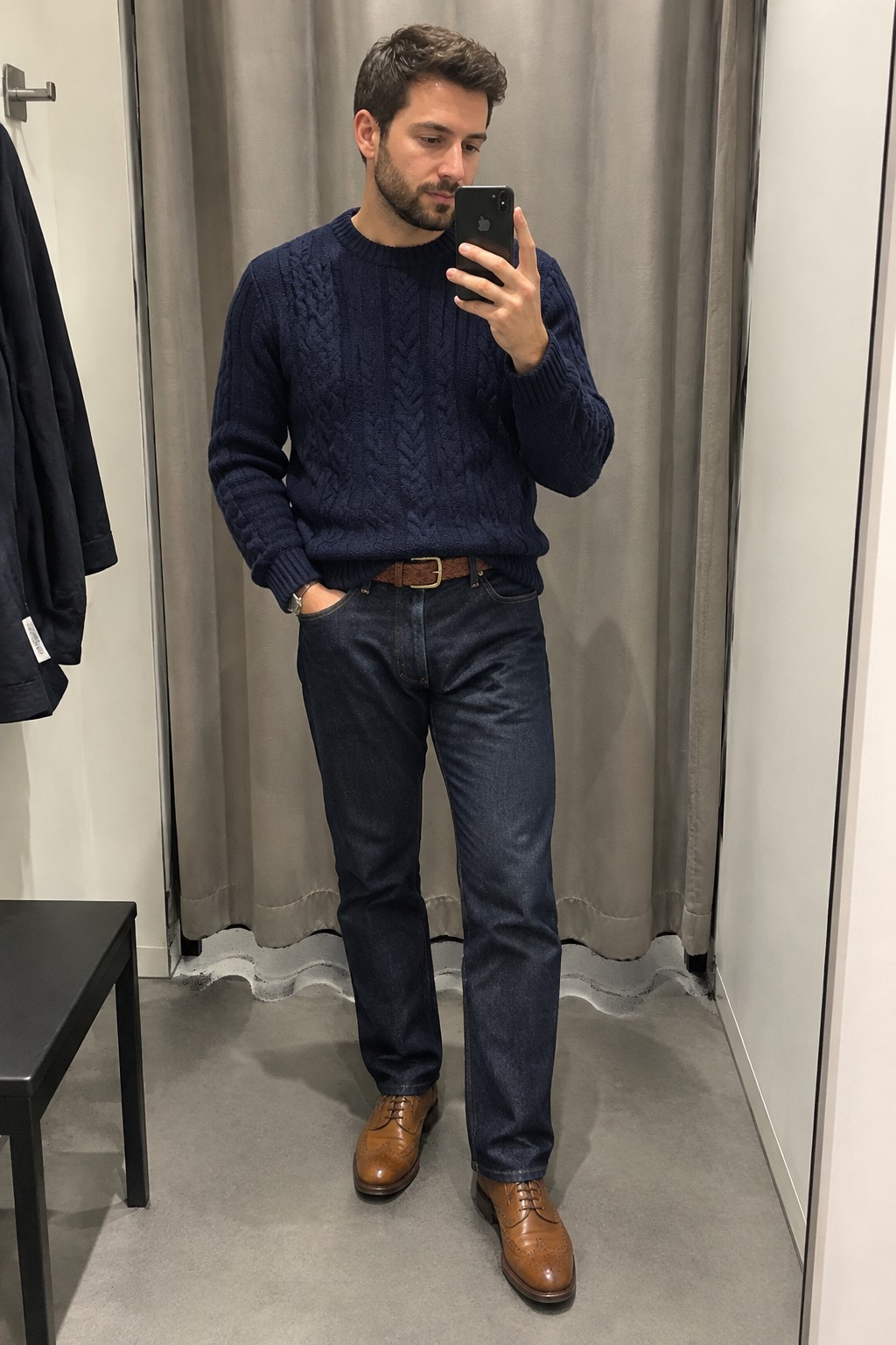A man wearing a navy cable-knit sweater, dark straight-leg jeans, a brown leather belt, and tan brogue shoes.