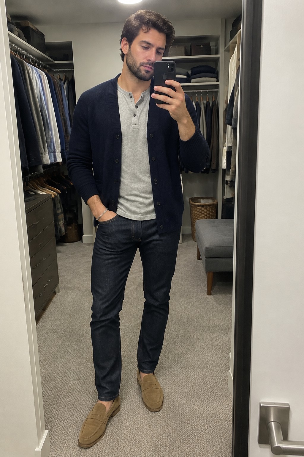 Man wearing a navy button-front cardigan over a gray henley shirt with dark jeans, tan suede loafers, and a silver bracelet.