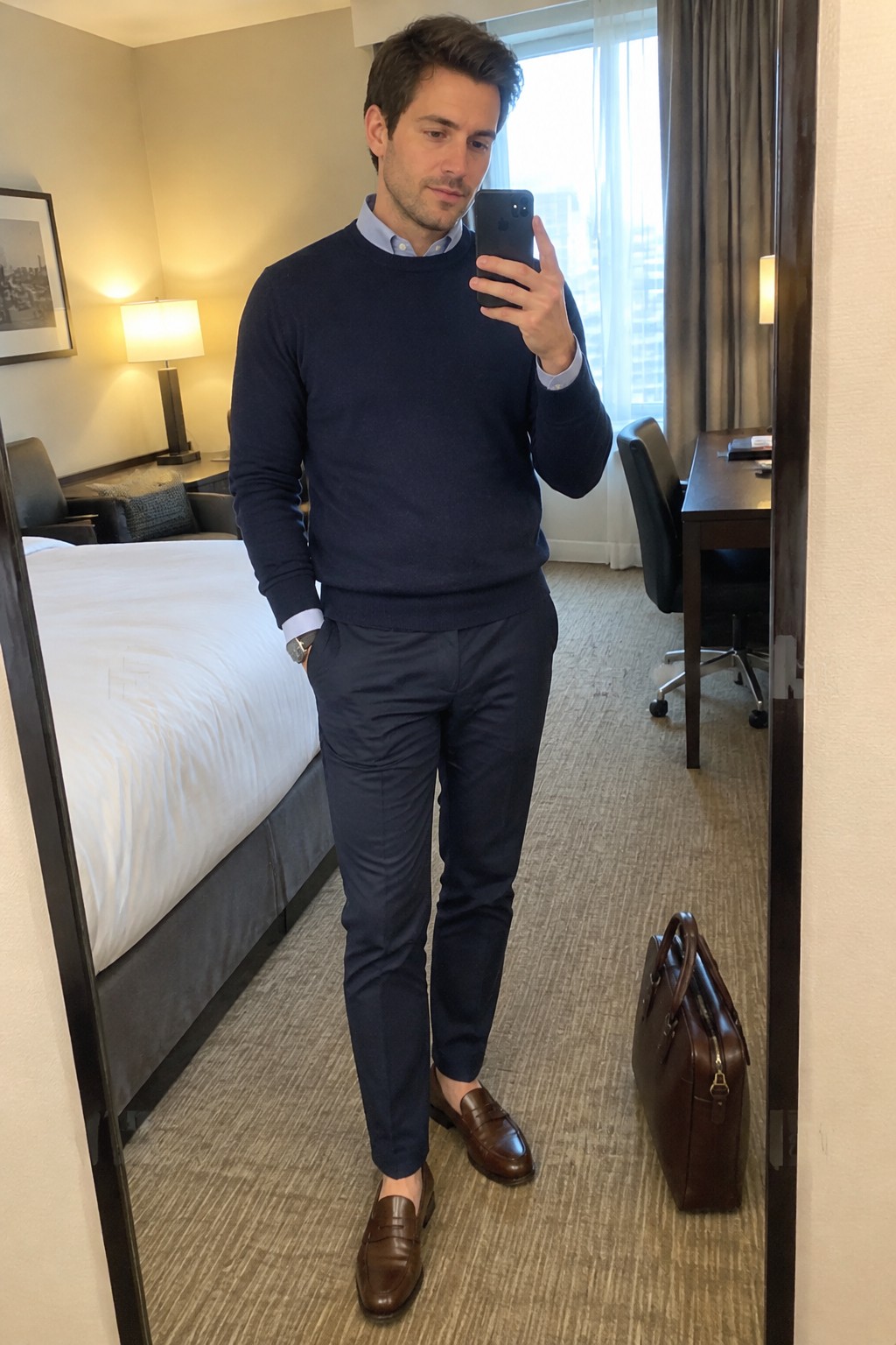Man wearing a navy crewneck sweater over a pale blue button-down shirt, dark tailored trousers, and brown leather loafers, holding a brown briefcase.