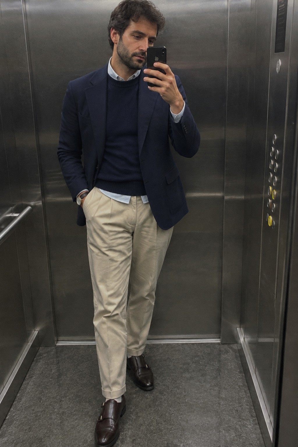 A man wearing a navy blazer over a navy sweater and light blue button-down shirt, with cream trousers, dark brown monk-strap shoes, and a wristwatch.