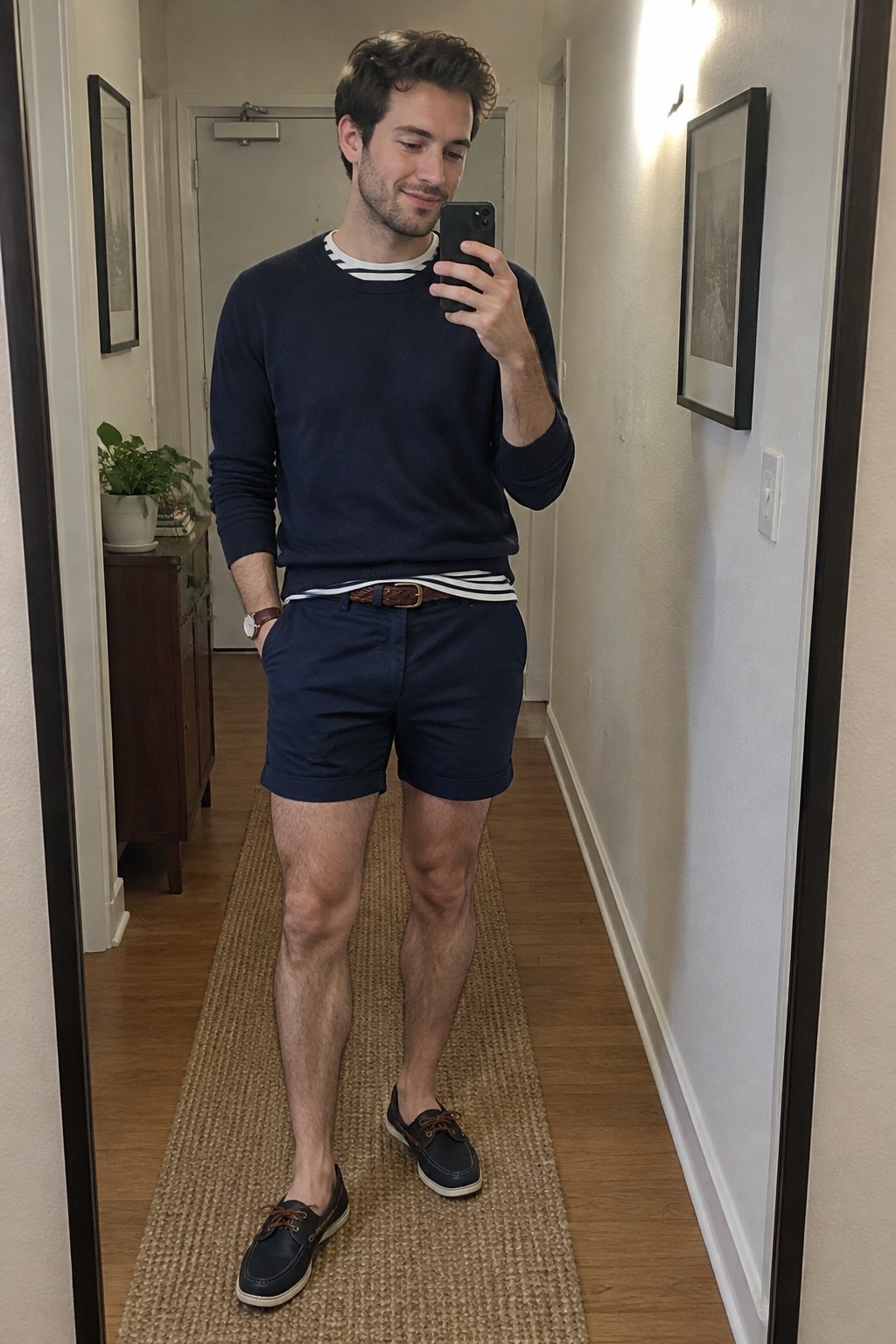 Man wearing a navy sweater over a black-and-white striped shirt, navy shorts with a brown belt, a wristwatch, and dark boat shoes with white soles.