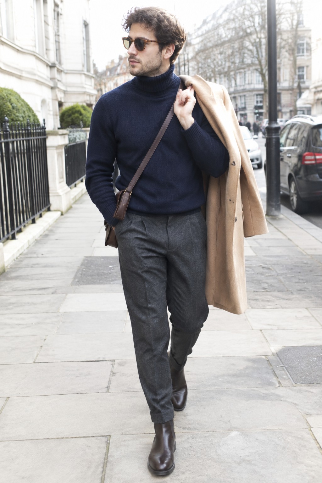 A man wears a navy ribbed turtleneck with charcoal gray trousers, a camel overcoat draped over one shoulder, brown leather ankle boots, sunglasses, and a crossbody bag.