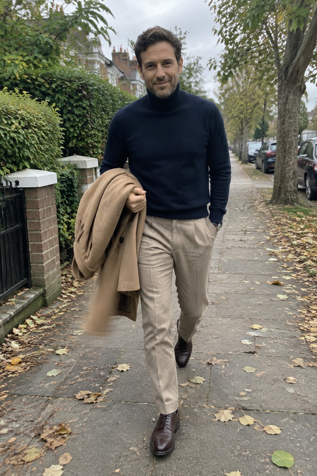 A man in a navy turtleneck, beige tailored trousers, and dark brown lace-up boots walks while carrying a camel coat.