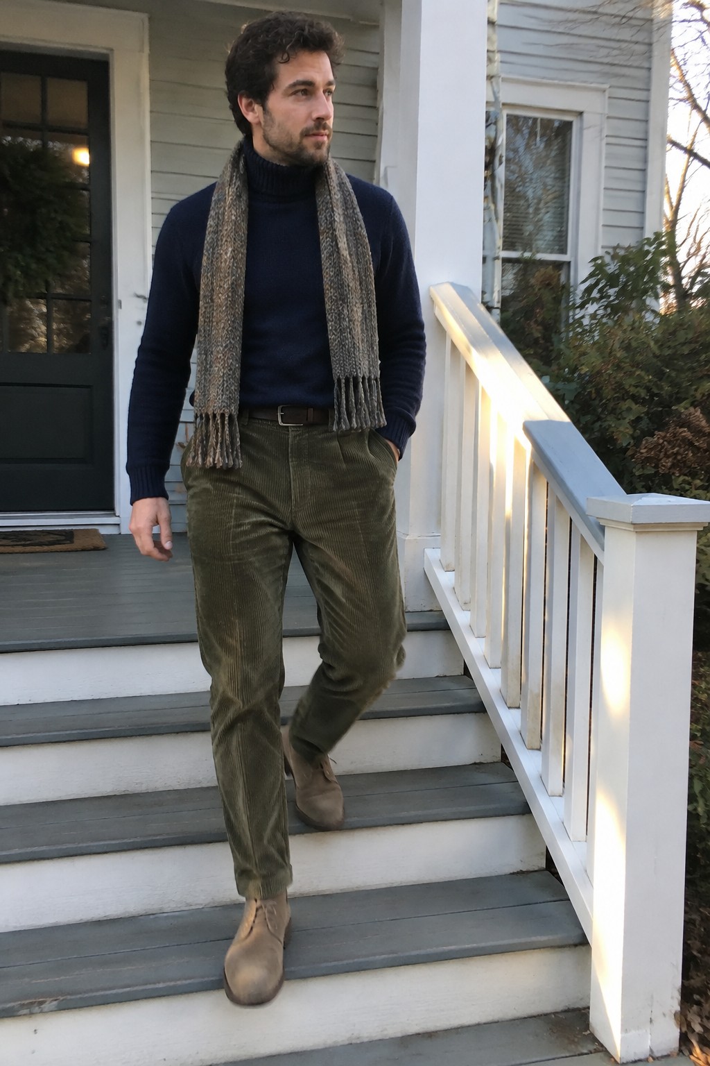 A man wearing a navy turtleneck, brown-gray scarf, olive corduroy trousers, dark belt, and taupe suede lace-up boots.