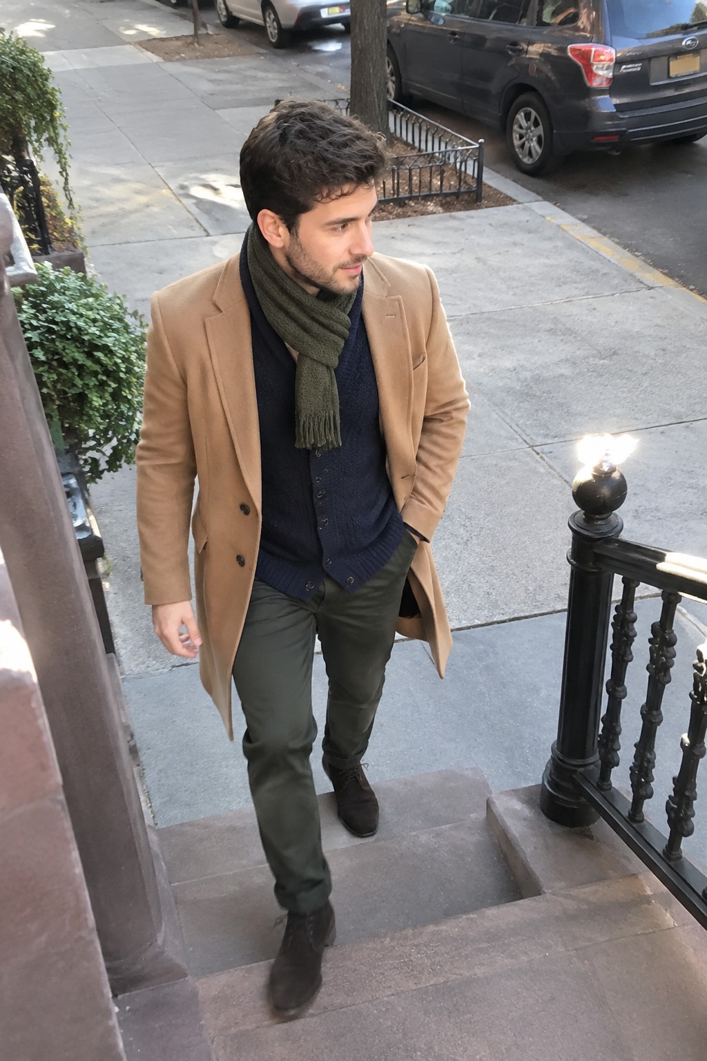 A man wearing a camel coat over a navy cardigan and dark sweater, an olive scarf, dark olive trousers, and brown lace-up boots.
