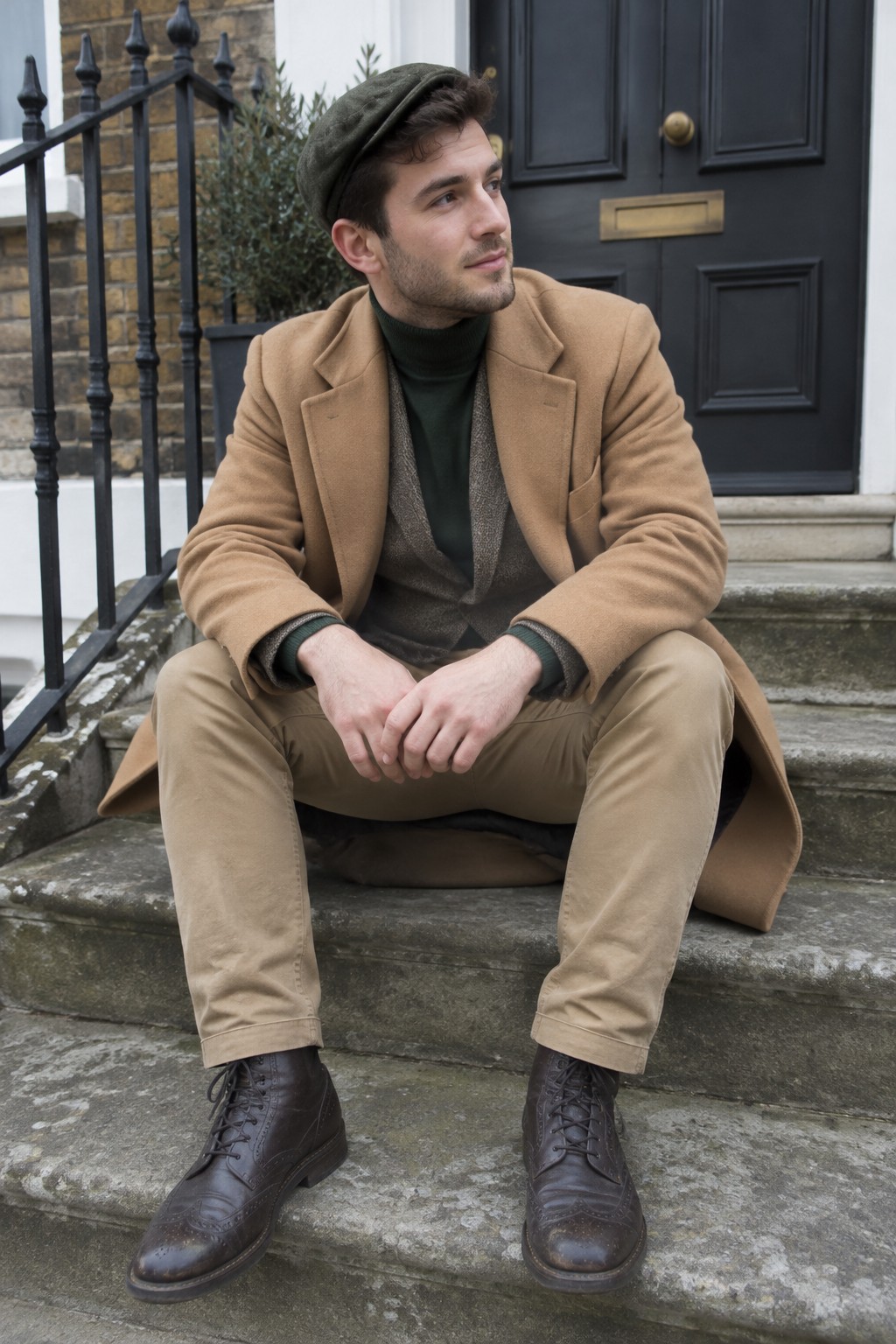 A man wearing a camel overcoat, dark green turtleneck, brown textured waistcoat, beige chinos, an olive cap, and dark brown brogue boots.