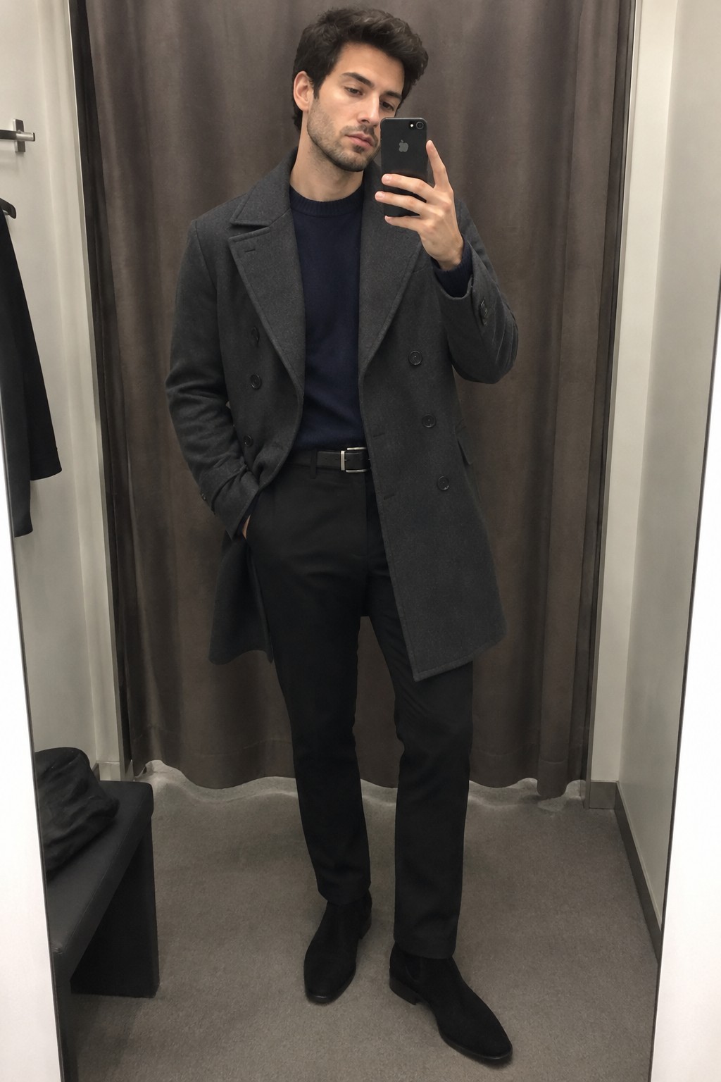 A man wearing a charcoal double-breasted wool coat over a navy crewneck sweater, black trousers, a black belt, and black ankle boots.
