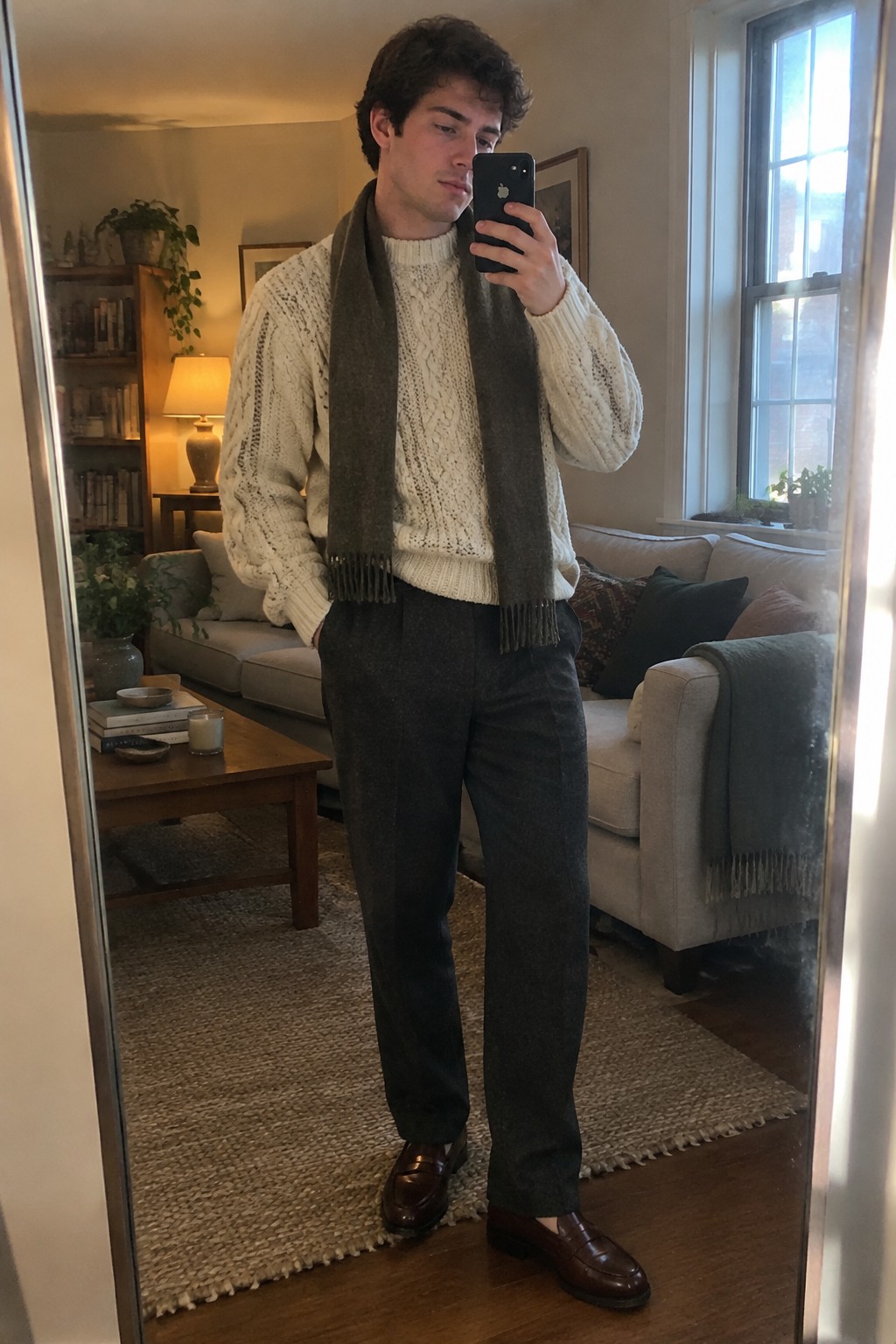 A man wears a cream cable-knit sweater, charcoal scarf, charcoal trousers, and brown leather loafers.