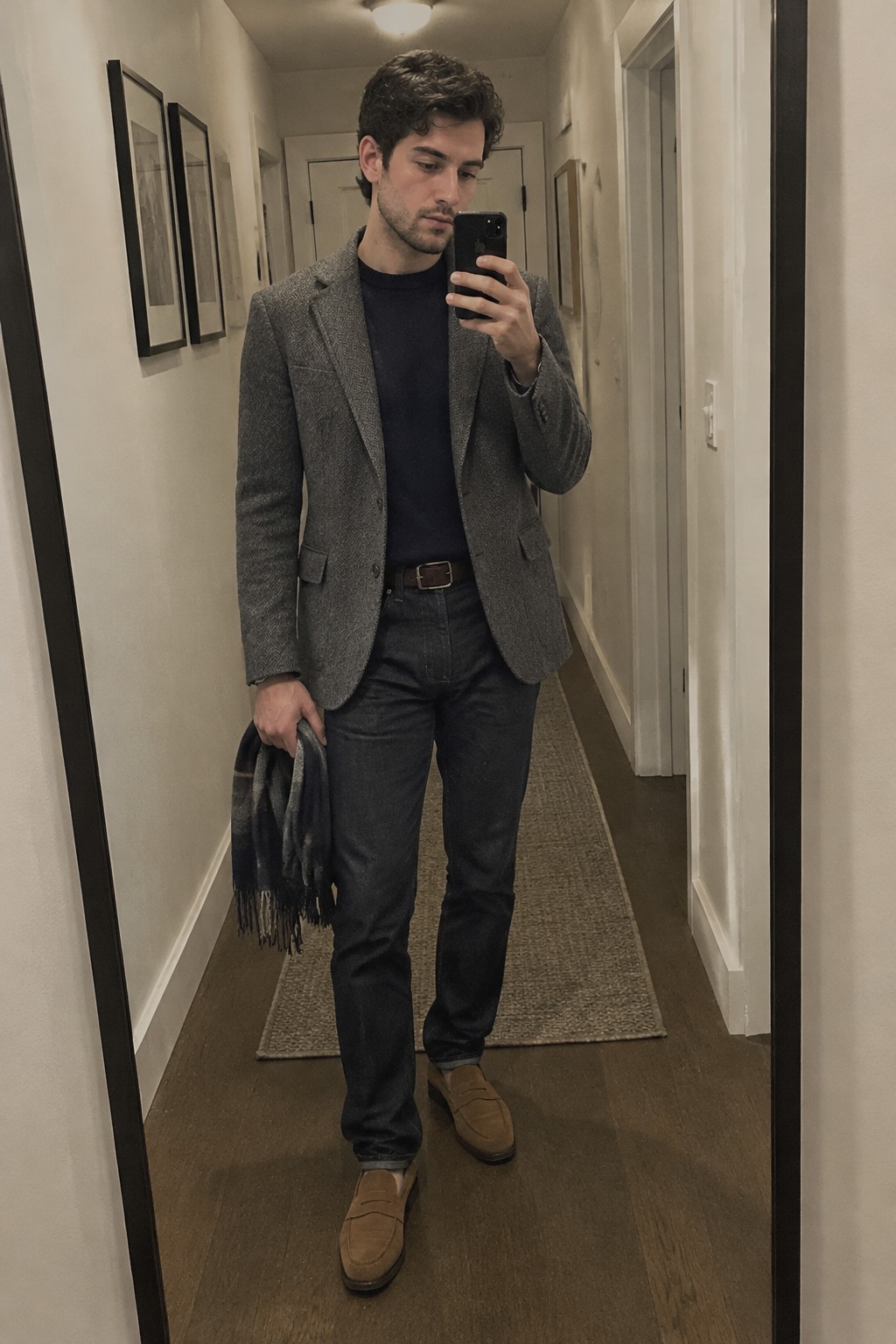 A man wears a grey tweed blazer over a navy sweater with dark jeans, a brown belt, tan suede loafers, and a charcoal fringed scarf.