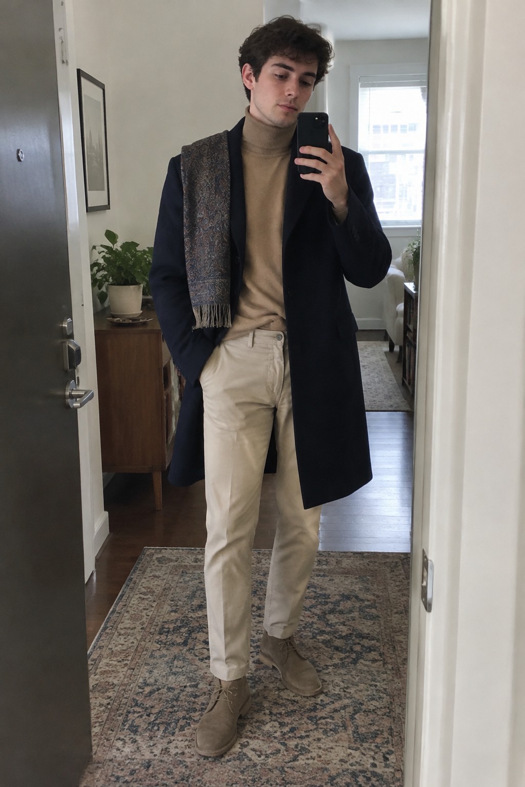 A man wearing a long navy overcoat, camel turtleneck, patterned scarf, cream trousers, and taupe suede lace-up shoes.