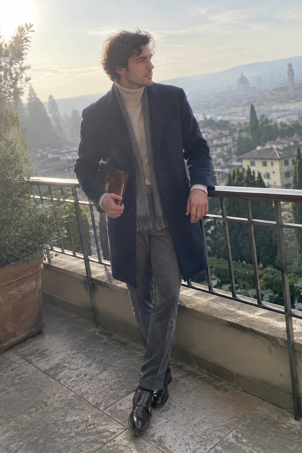 A man wearing a navy overcoat, cream ribbed turtleneck, grey scarf and trousers, dark leather monk-strap shoes, and carrying a brown leather folder.