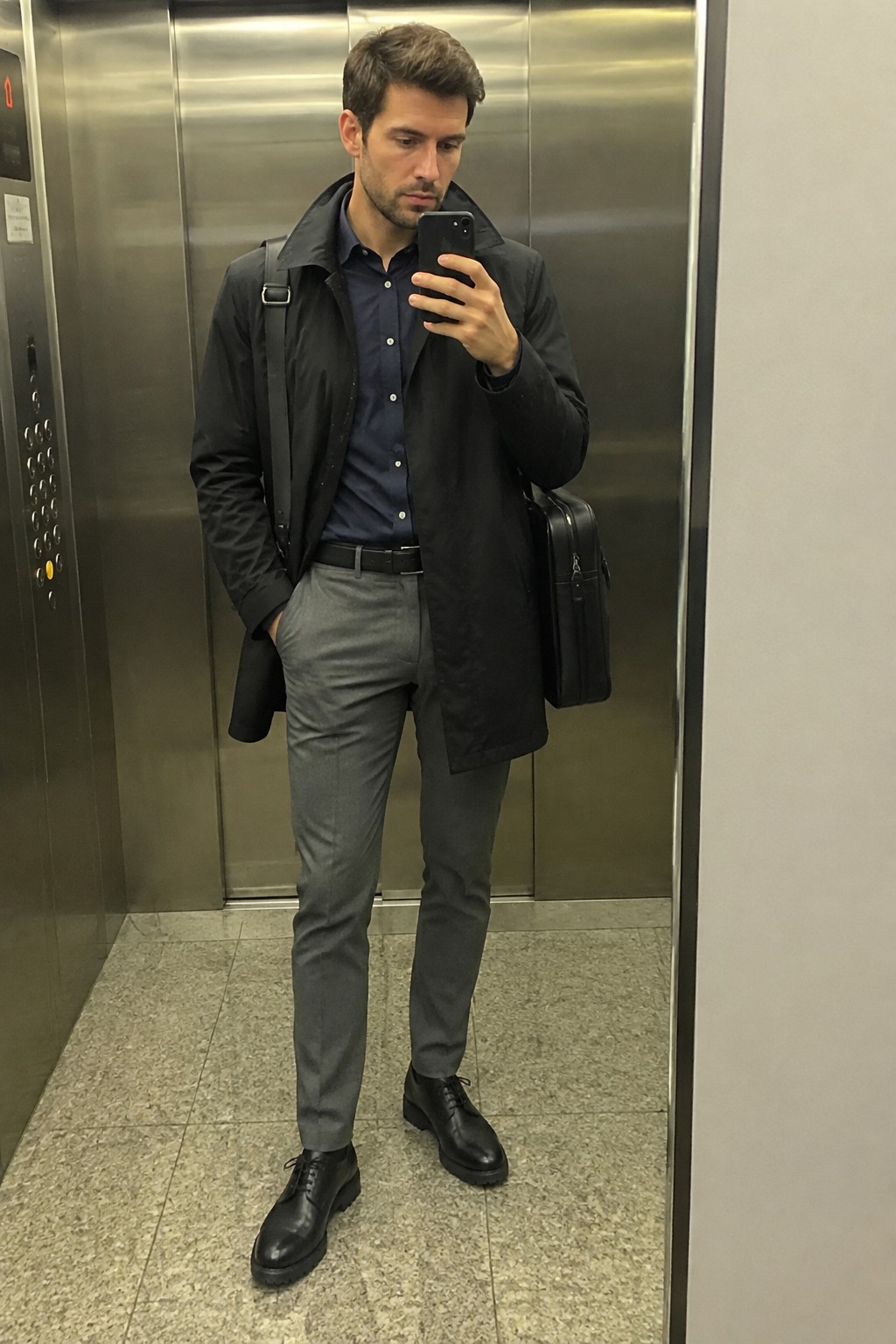 Man wearing a black raincoat over a navy button-down shirt with gray tailored trousers, black belt, black lace-up shoes, and a shoulder bag.