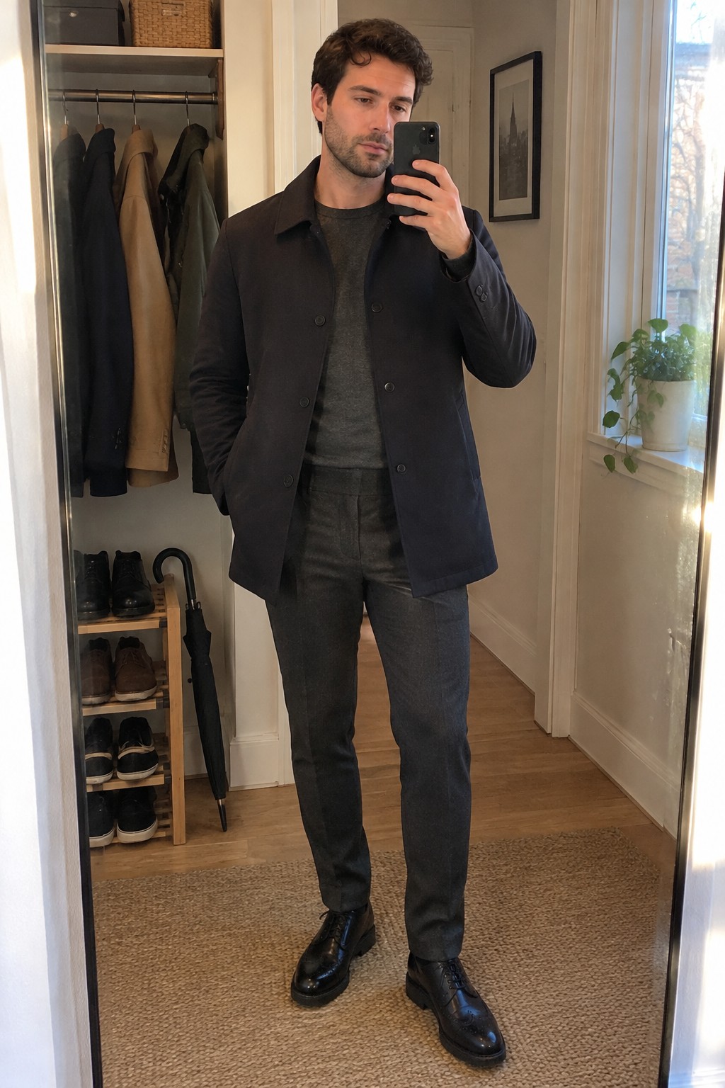 A man wears a dark buttoned coat over a charcoal crew-neck sweater and matching tailored trousers with black lace-up shoes.
