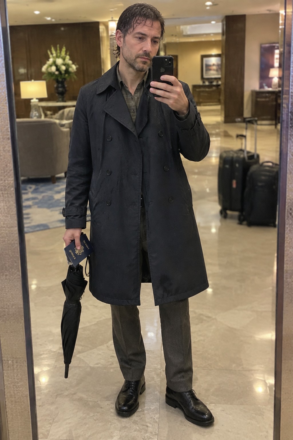 A man wears a long dark double-breasted coat over a muted checked shirt and charcoal trousers with black shoes, holding a folded umbrella and passport.