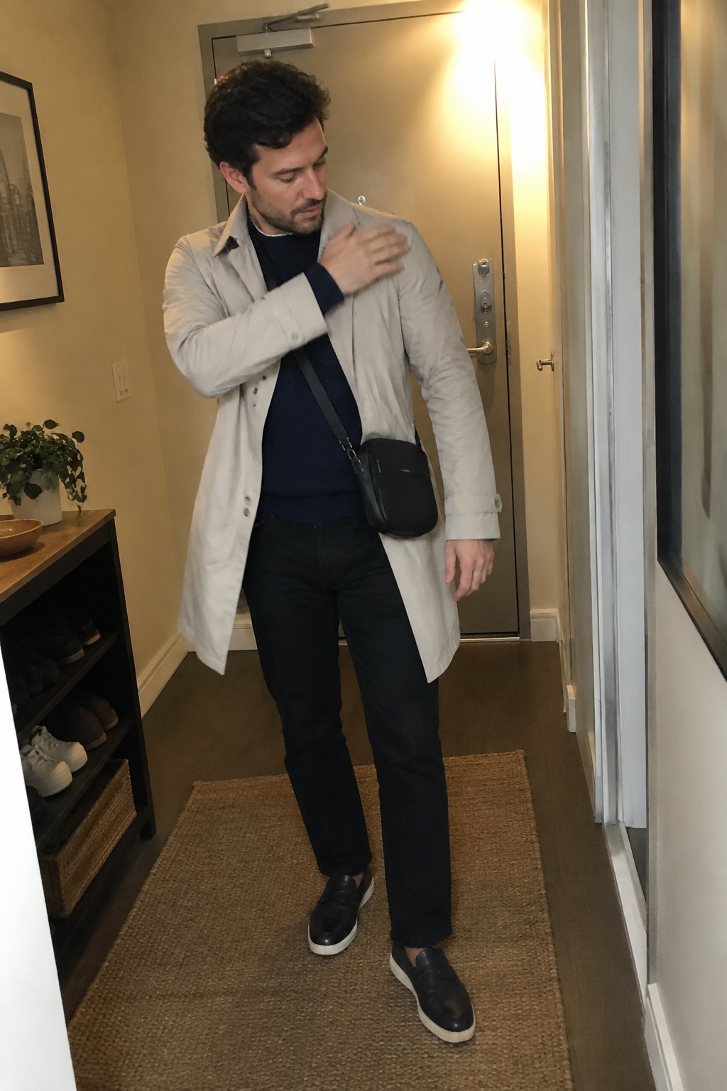 A man wears a light gray raincoat over a navy sweater with black jeans, black slip-on shoes with white soles, and a black crossbody bag.