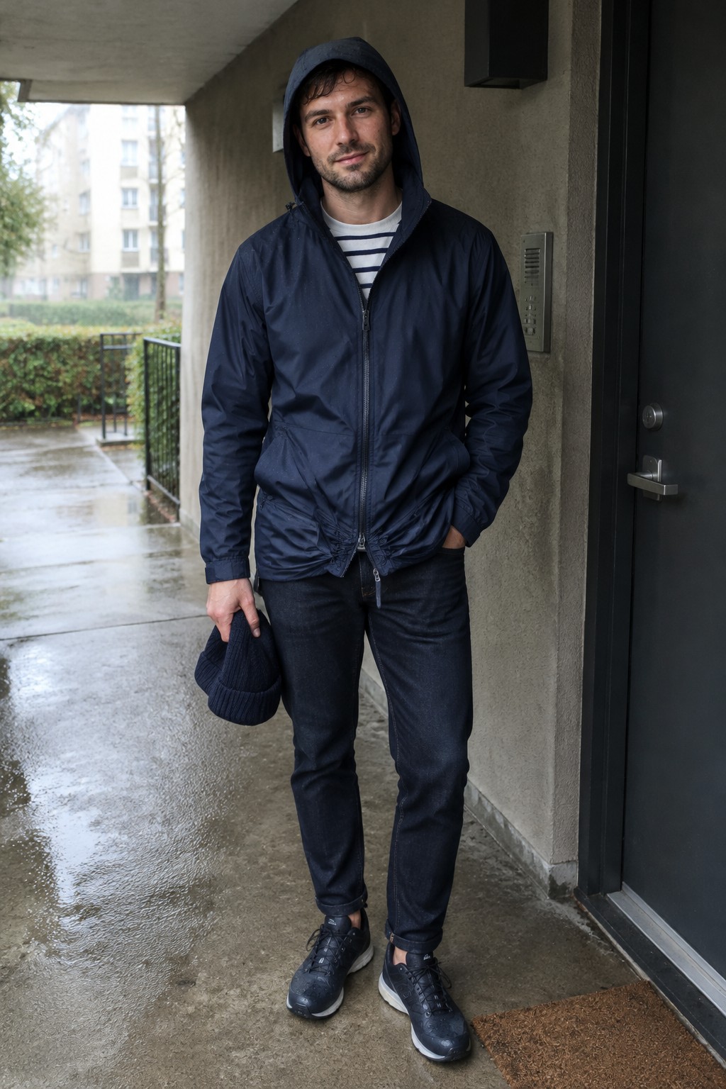 A man wearing a navy hooded rain jacket over a navy-and-white striped shirt, dark jeans, and dark sneakers, holding a navy beanie.