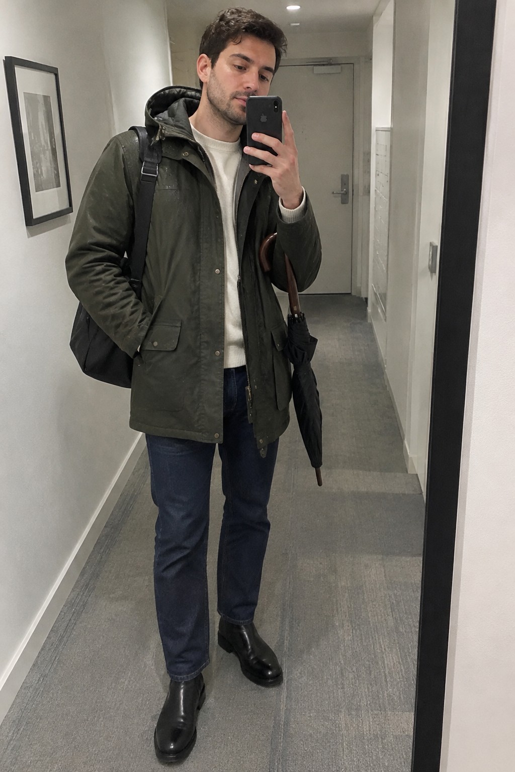 A man wears a dark olive hooded field jacket over a cream sweater, dark jeans, and black Chelsea boots, carrying a black backpack and umbrella.
