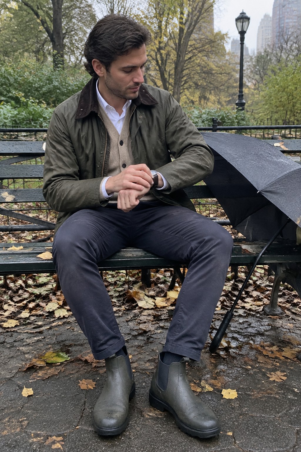 A man in an olive waxed jacket with a corduroy collar, taupe cardigan, white shirt, charcoal trousers, and gray rubber Chelsea boots.