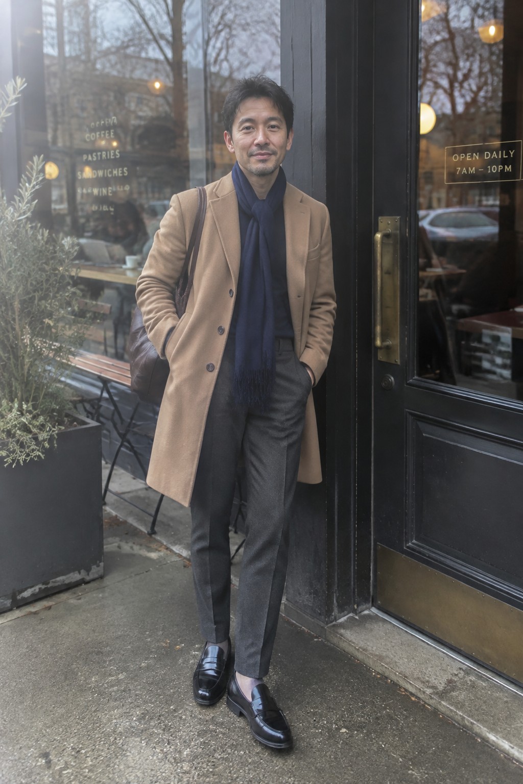 A man wearing a camel overcoat, navy scarf, gray high-rise trousers, black leather loafers, and a brown shoulder bag.