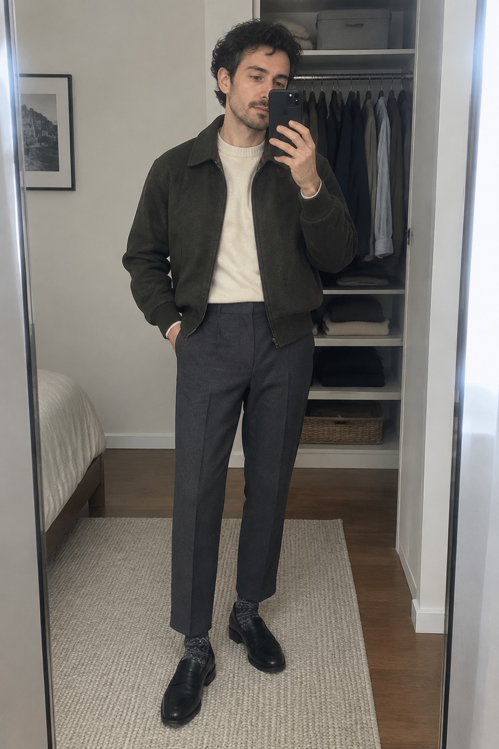 A man wears a dark green zip-front jacket over a cream sweater, charcoal high-rise trousers, patterned socks, and black loafers.