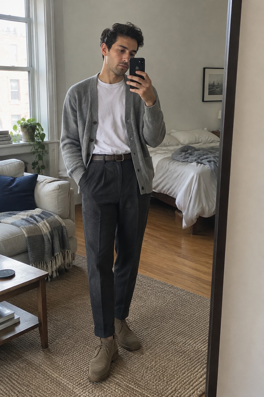 A man wears a gray cardigan over a white T-shirt, charcoal high-rise trousers with turned-up cuffs, a dark belt, and taupe lace-up boots.