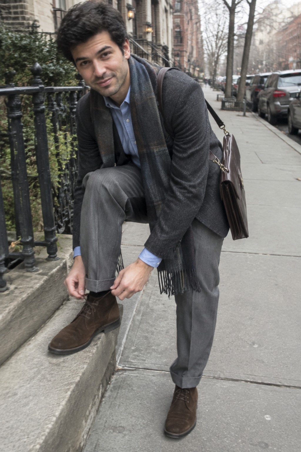 A man wearing high-rise gray cuffed trousers, brown suede ankle boots, a light blue shirt, a charcoal cardigan and scarf, and a brown leather shoulder bag.