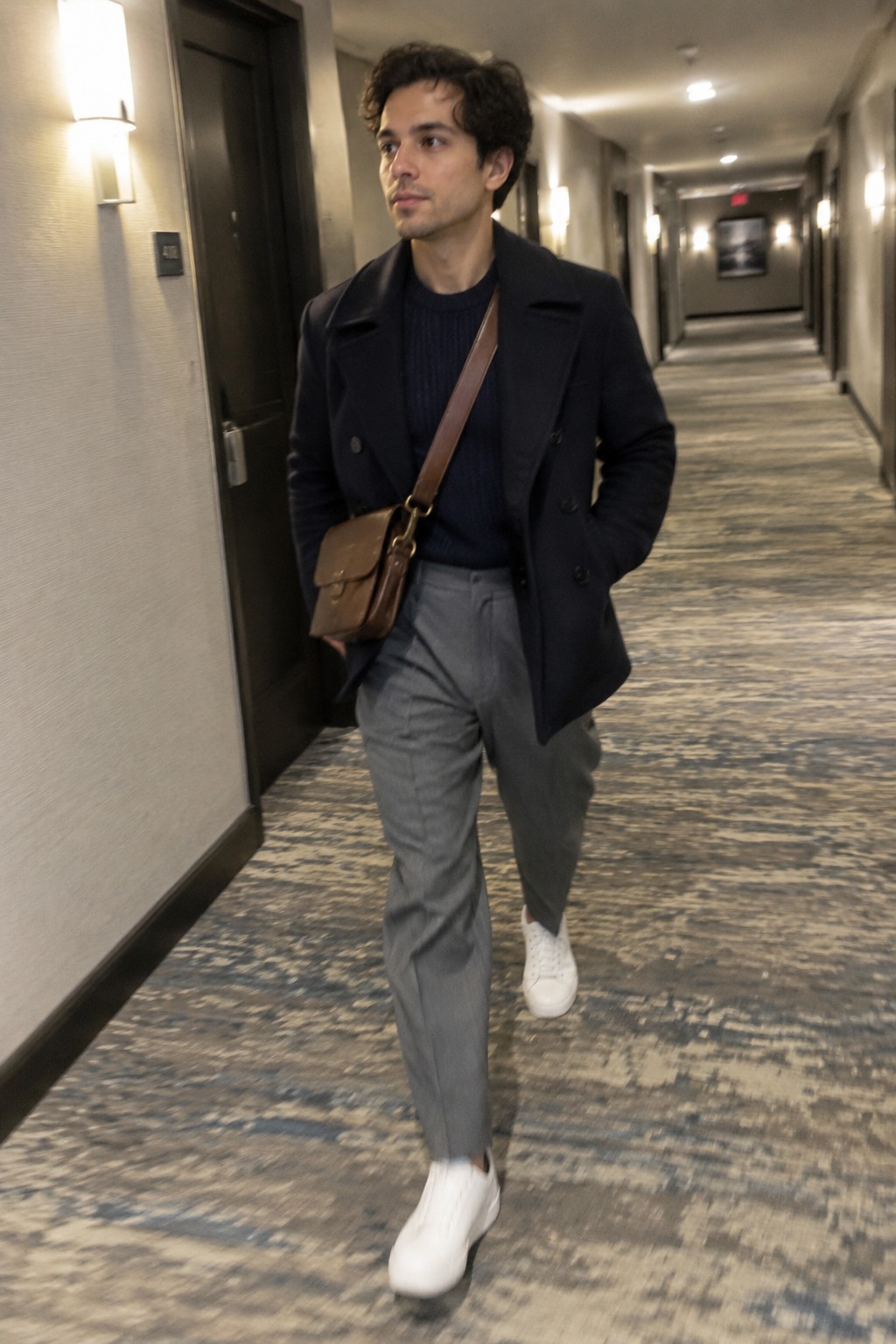 A man wears a navy double-breasted peacoat over a navy sweater, gray high-rise trousers, white sneakers, and a brown leather crossbody bag.