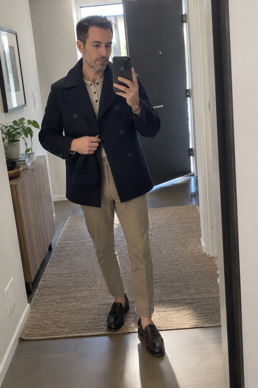Man wearing a navy double-breasted peacoat over a cream henley, beige high-rise tapered trousers, and dark brown loafers.