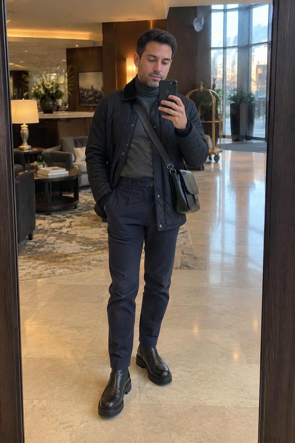 A man wears a black quilted jacket over a charcoal turtleneck, navy high-rise trousers, black Chelsea boots, and a black crossbody bag.