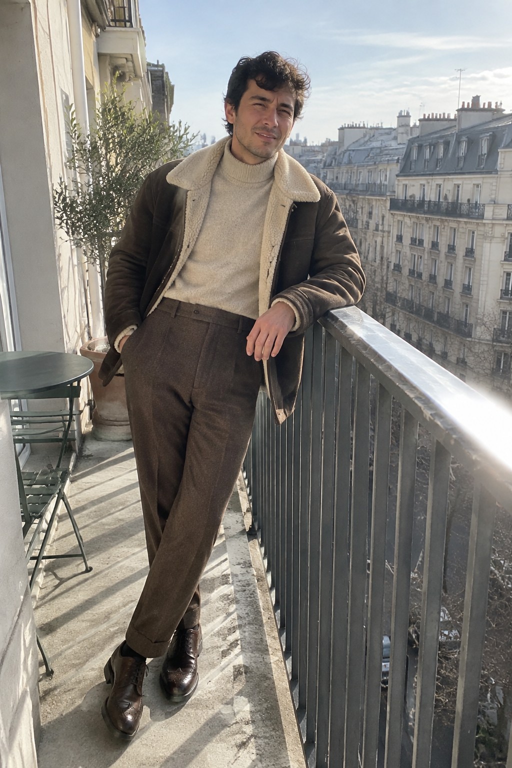 A man wearing a dark brown shearling-lined jacket, cream turtleneck sweater, high-rise brown trousers, and brown leather lace-up shoes.