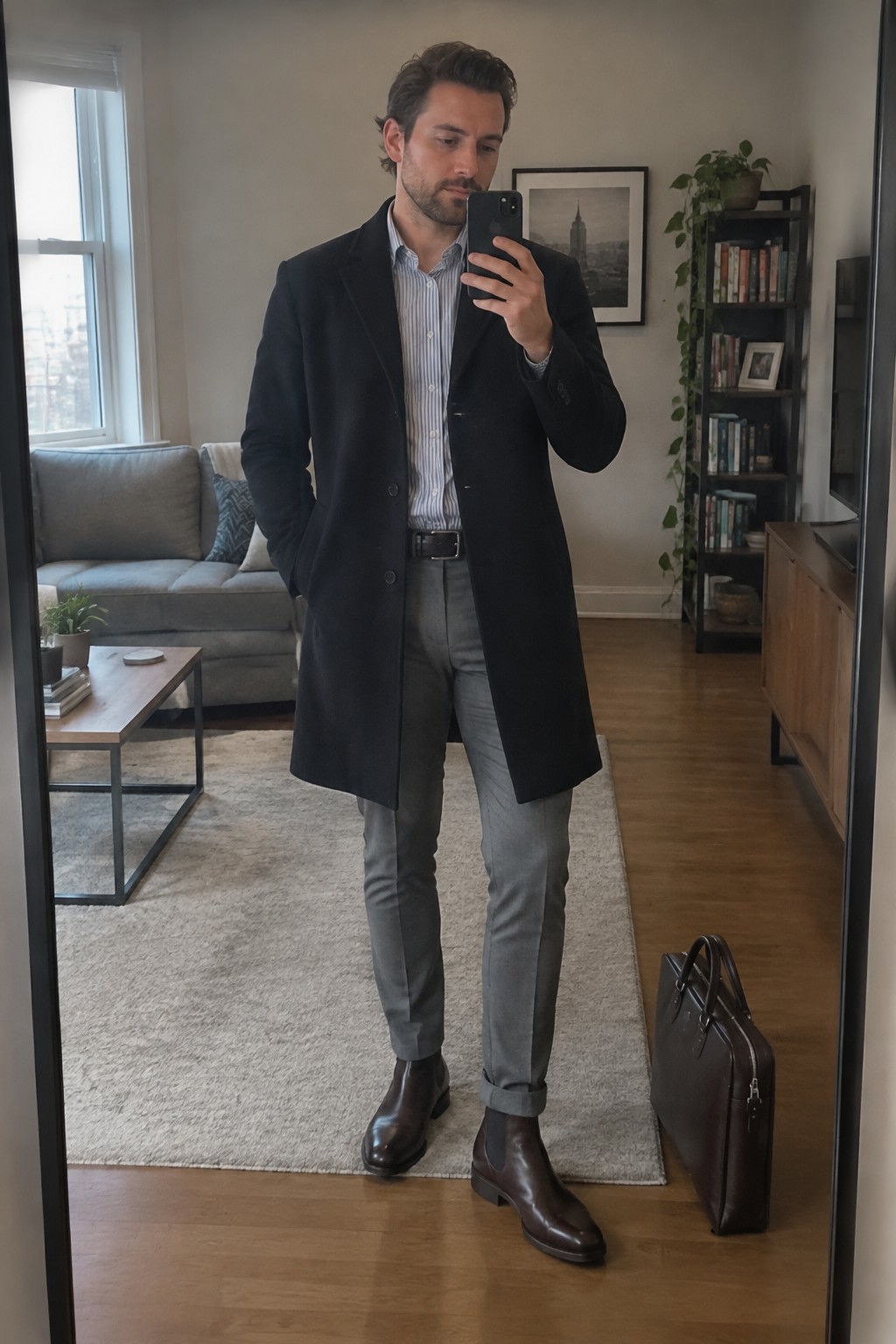 A man wearing a long black overcoat over a blue-and-white striped shirt, gray trousers, brown Chelsea boots, and carrying a dark leather briefcase.