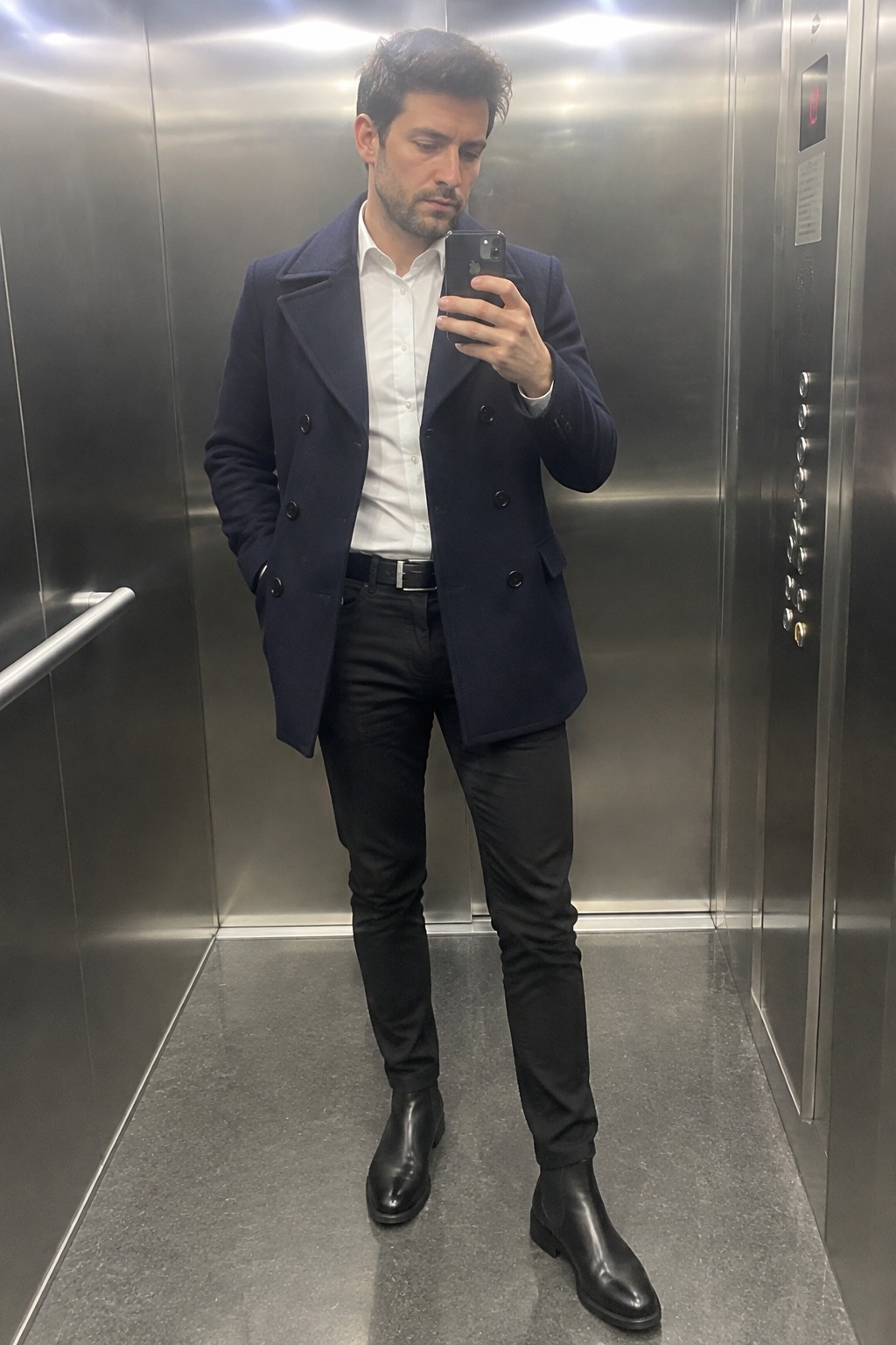 Man wearing a navy double-breasted peacoat over a white button-down shirt, black trousers, a black belt, and black leather Chelsea boots.