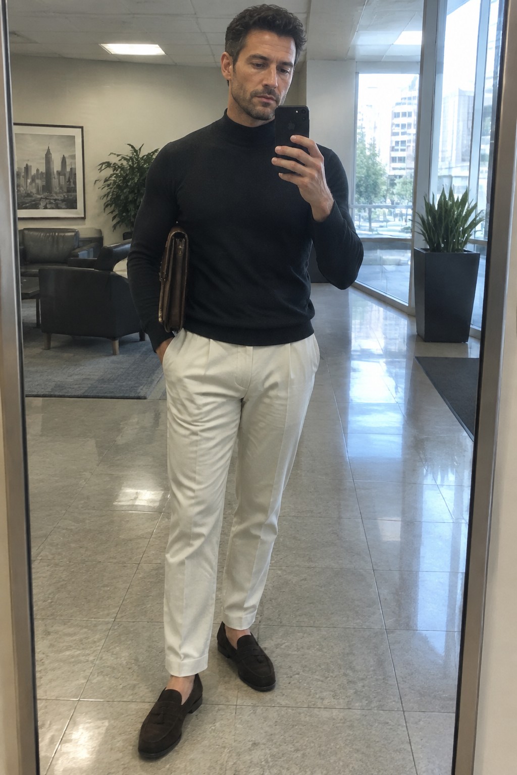 A man taking a mirror selfie in a black turtleneck, cream trousers, dark brown loafers, and carrying a slim leather folio.