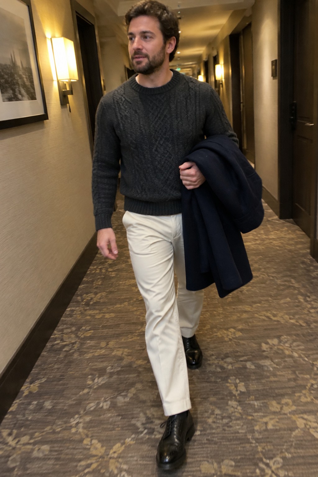 A man wearing a charcoal cable-knit crewneck sweater, cream straight-leg trousers, black leather ankle boots, and carrying a navy coat.