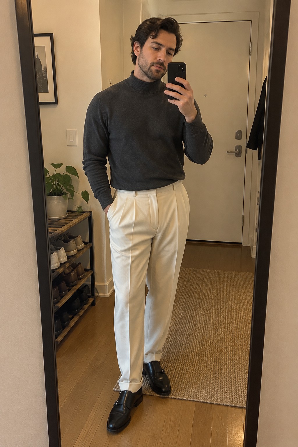 A man wearing a charcoal ribbed turtleneck tucked into pleated cream trousers with cuffed hems and black leather loafers.