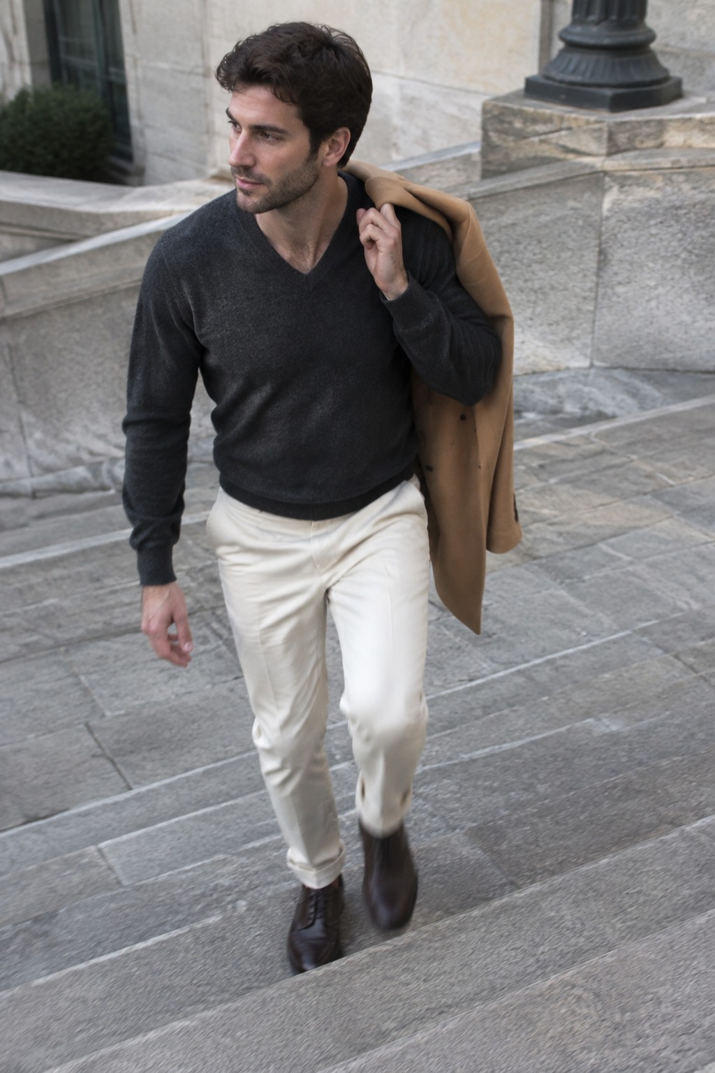 A man wearing a charcoal V-neck sweater, cream trousers, dark leather shoes, and a camel coat draped over one shoulder.