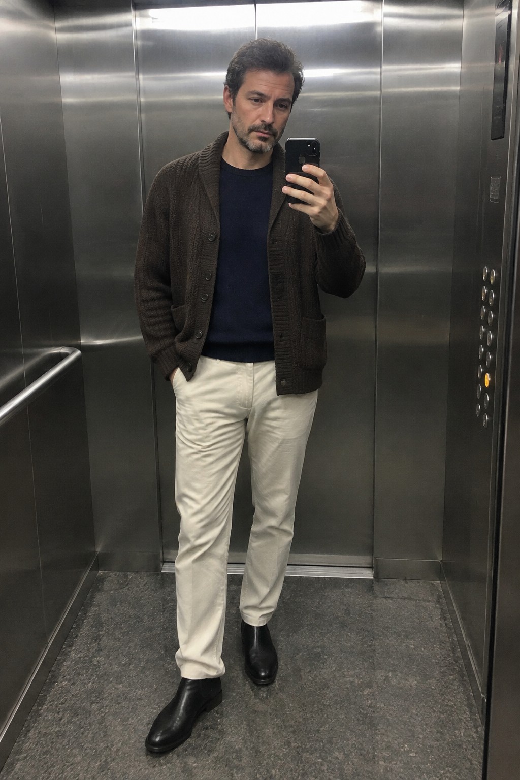 A man wears cream trousers, a navy crewneck sweater, a dark brown cardigan, and black leather ankle boots.