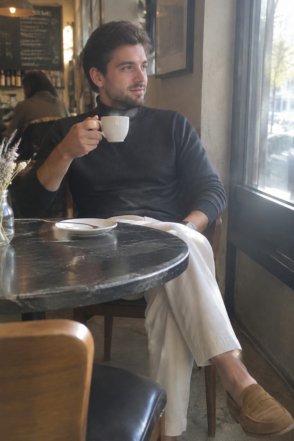 A man wearing a charcoal turtleneck, cream cropped trousers, and brown suede loafers sits at a café table holding a white cup.