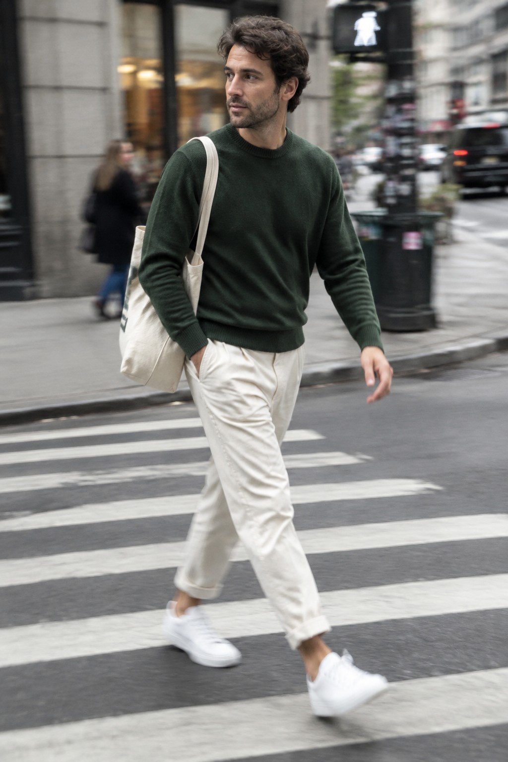 A man walks in a forest green crewneck sweater, relaxed cream trousers, white sneakers, and a cream canvas tote.