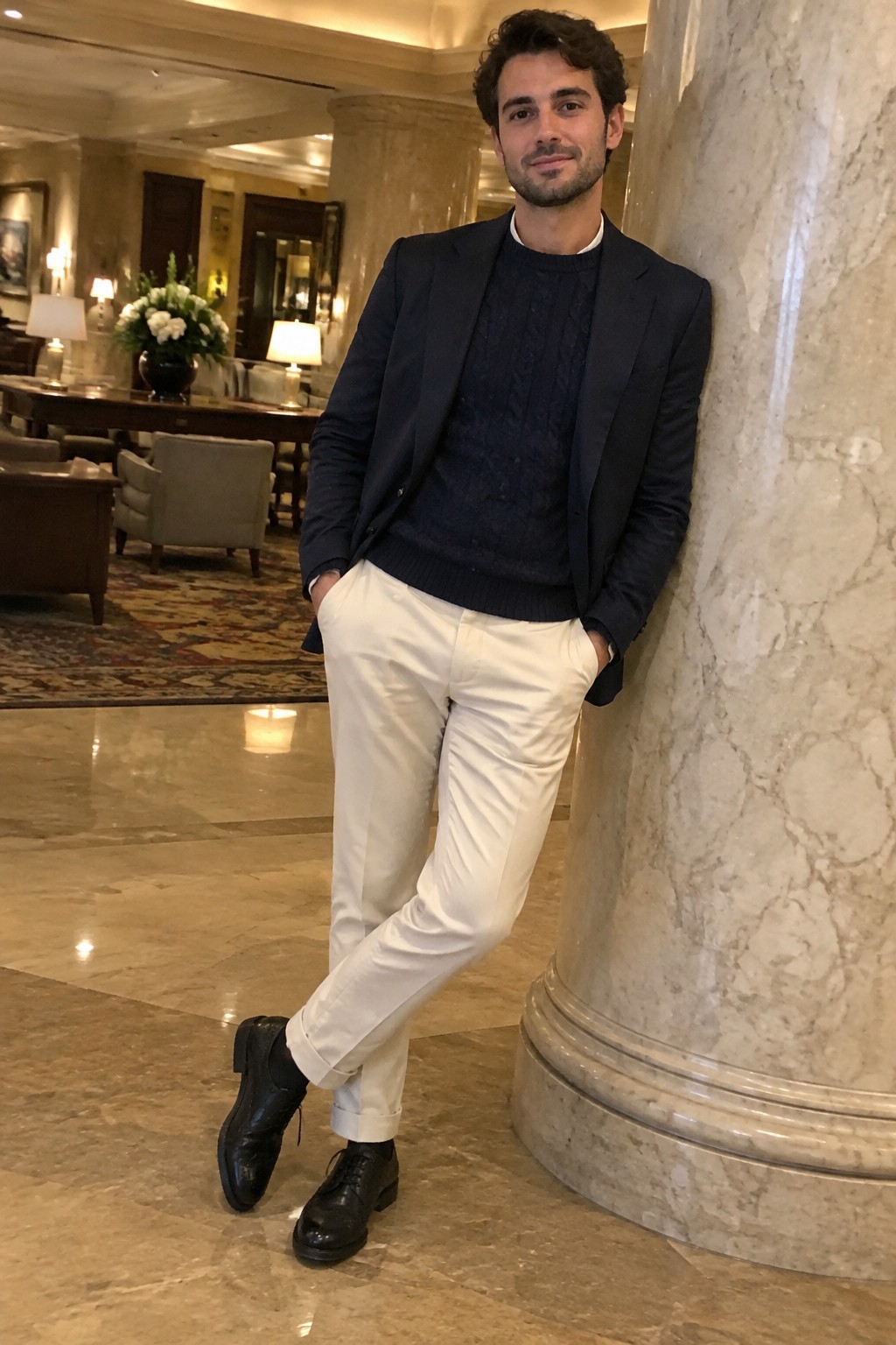A man wearing a navy cable-knit sweater and dark blazer with cream cuffed trousers and black leather lace-up shoes.