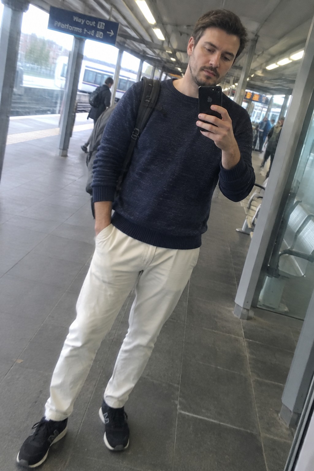 Man wearing a navy crewneck sweater, cream trousers, black sneakers, and a black backpack.