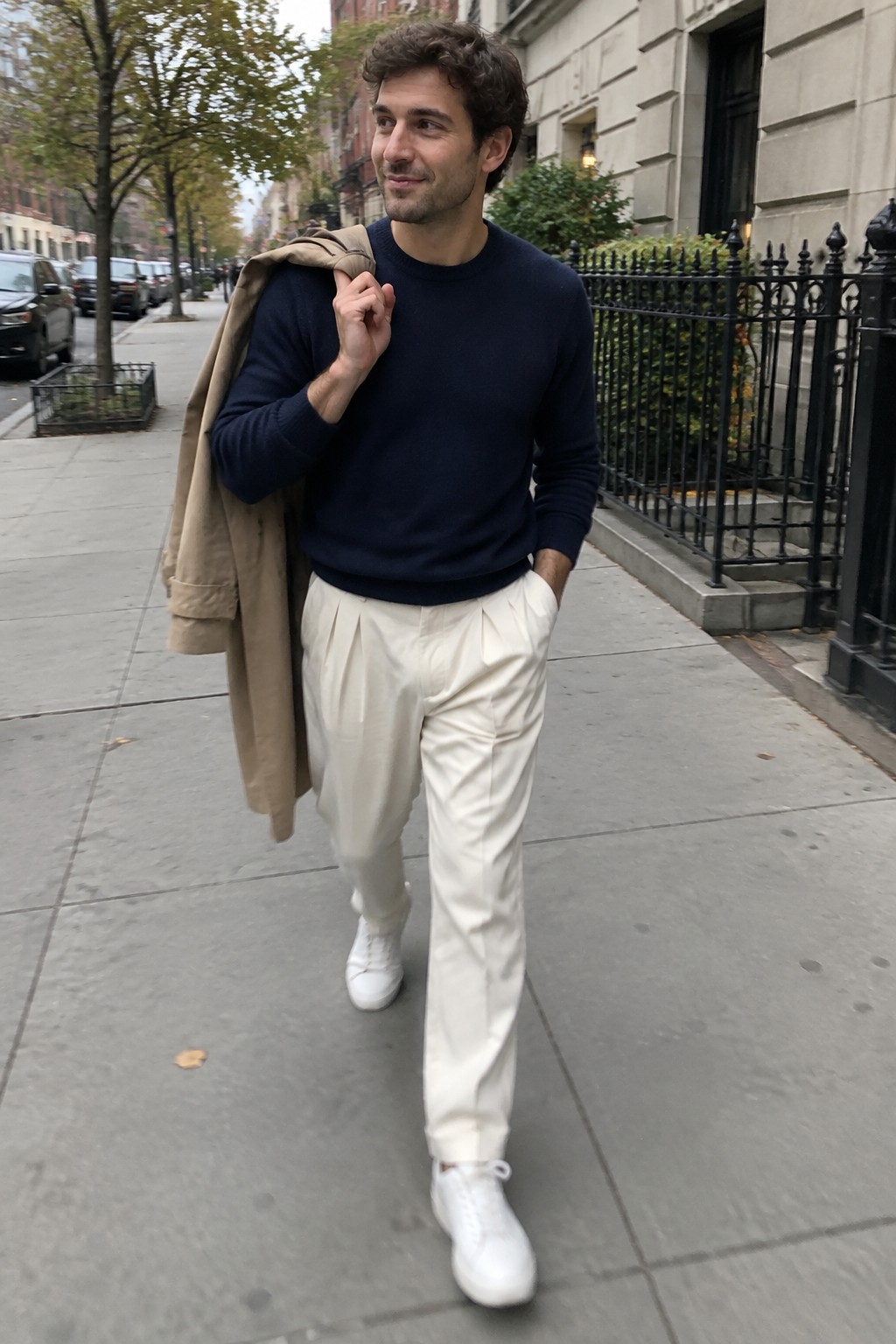 A man wearing a navy crewneck sweater, cream pleated trousers, white sneakers, and a tan coat draped over his shoulder.