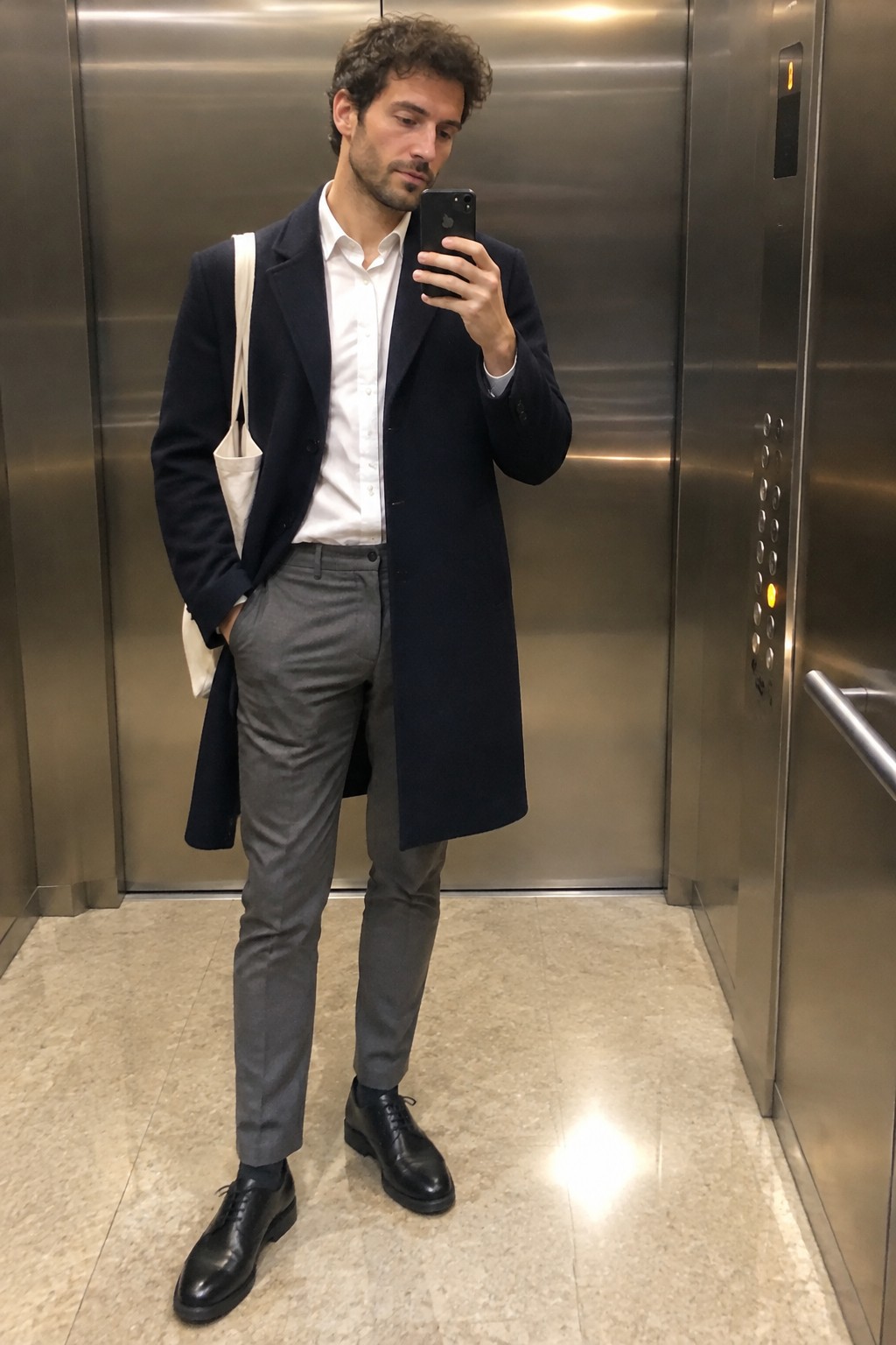 A man wearing a long navy wool overcoat, white button-down shirt, gray trousers, black lace-up shoes, and a cream canvas tote.