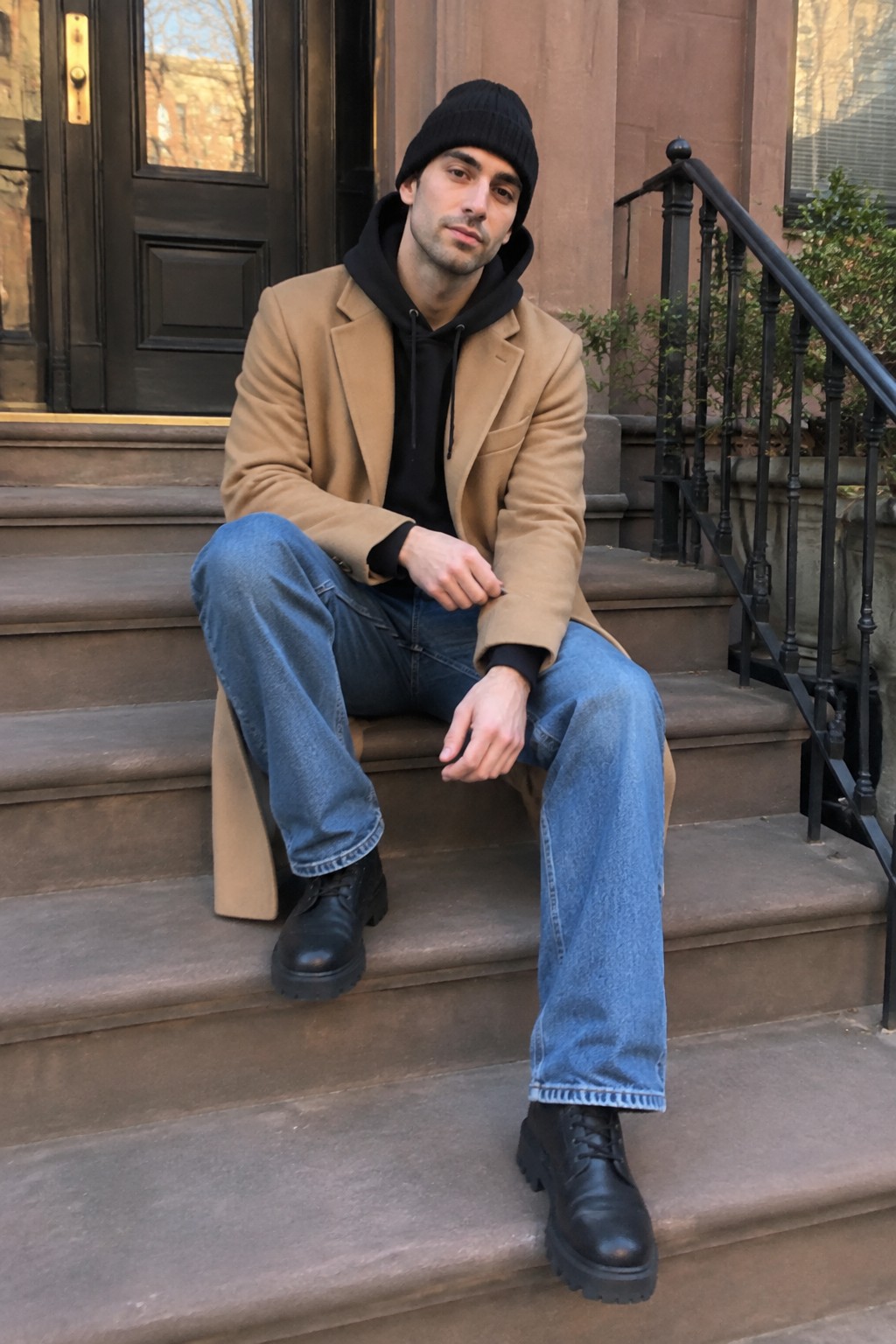 A man wearing a camel longline overcoat over a black hoodie, with a black knit beanie, loose blue jeans, and black lace-up boots, seated on stone steps.